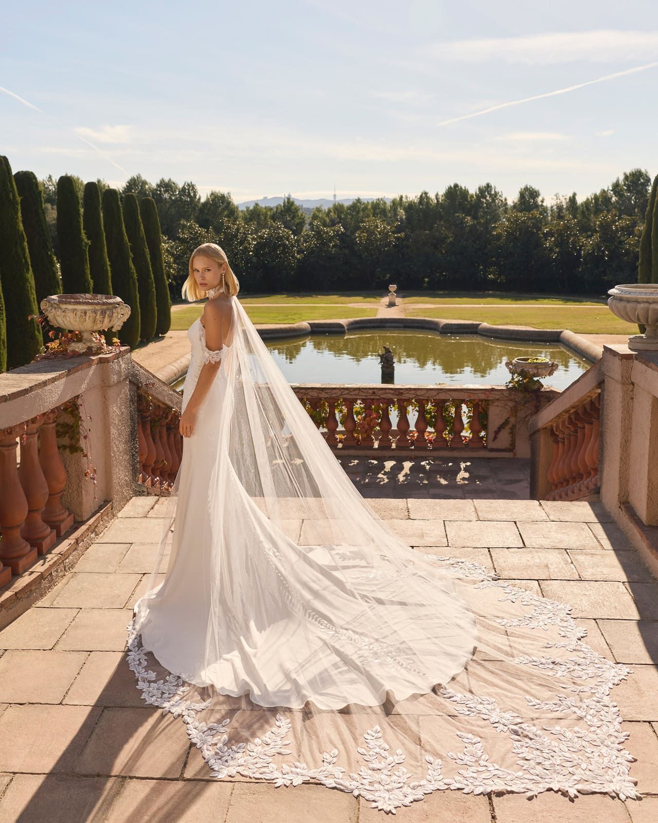 Novia con vestido blanco y largo velo de encaje, de pie en una terraza junto a un jardín formal y un estanque.