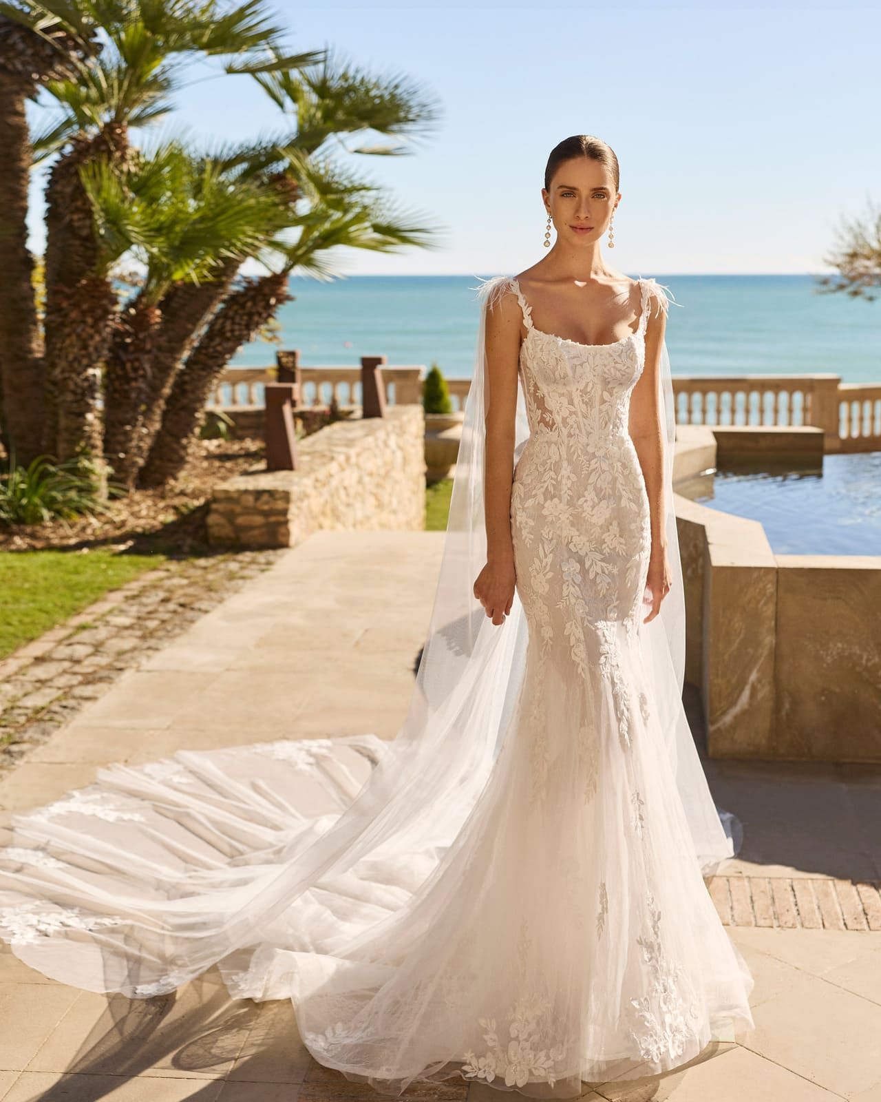 Mujer con un vestido de novia de encaje blanco de pie en un patio junto al mar con palmeras y el océano al fondo.