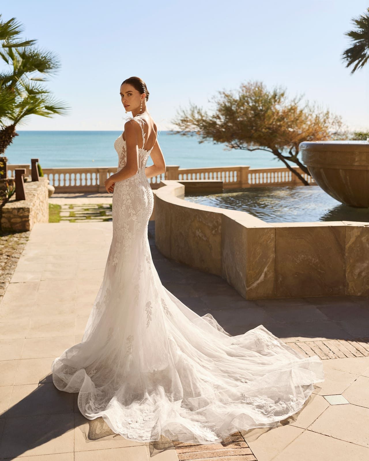 Novia con un vestido de encaje blanco de pie junto a una terraza junto al mar con vistas al océano y una fuente.