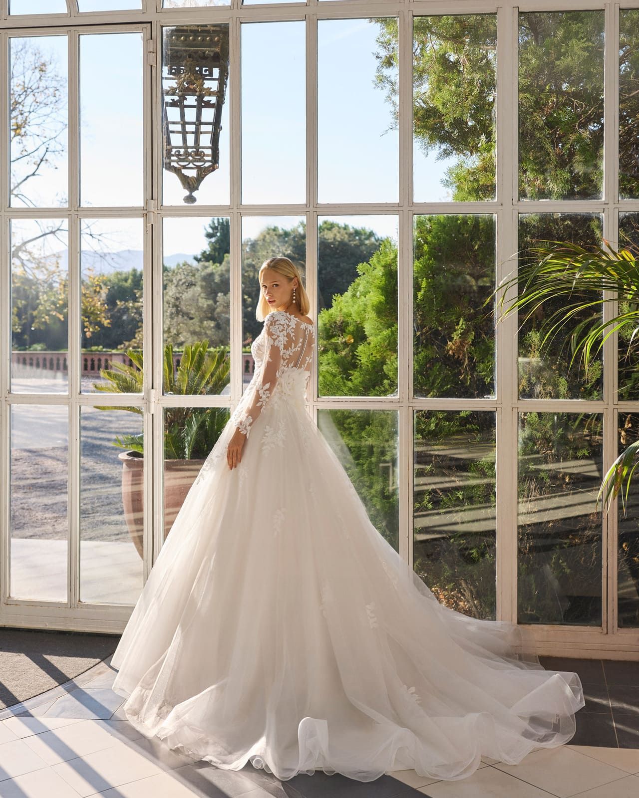 Novia con vestido de novia blanco de pie junto a grandes ventanales en una habitación iluminada por el sol.