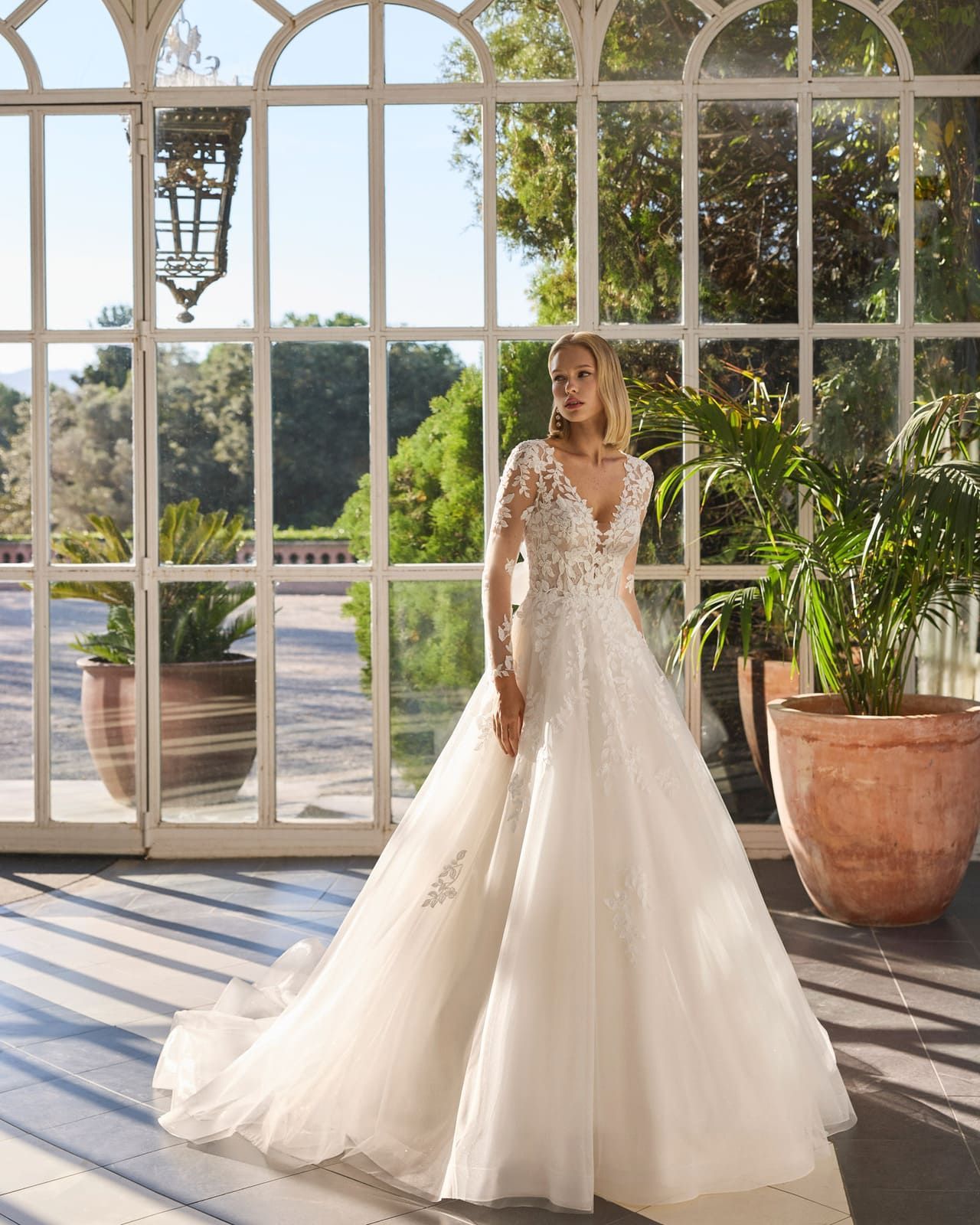 Novia con vestido blanco de pie frente a altos ventanales arqueados en un luminoso jardín acristalado.