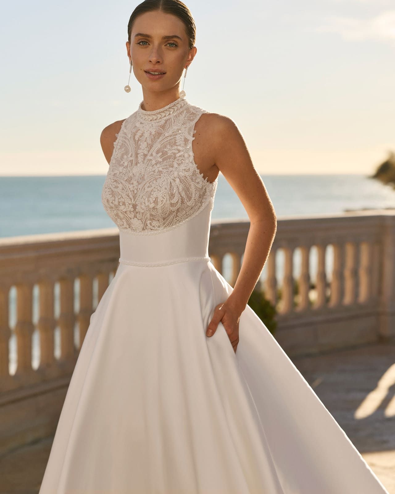 Mujer con un vestido blanco de encaje con escote halter de pie en una terraza junto al mar al atardecer.