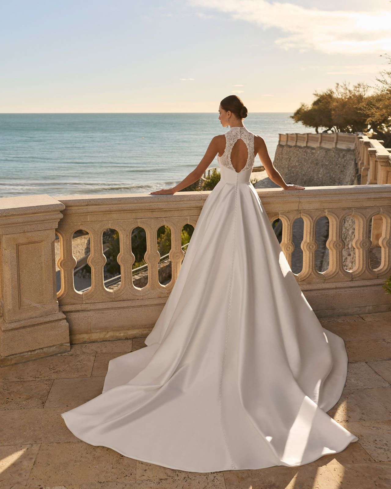 Novia con vestido blanco de pie en una terraza junto al mar al atardecer, mirando hacia el océano.