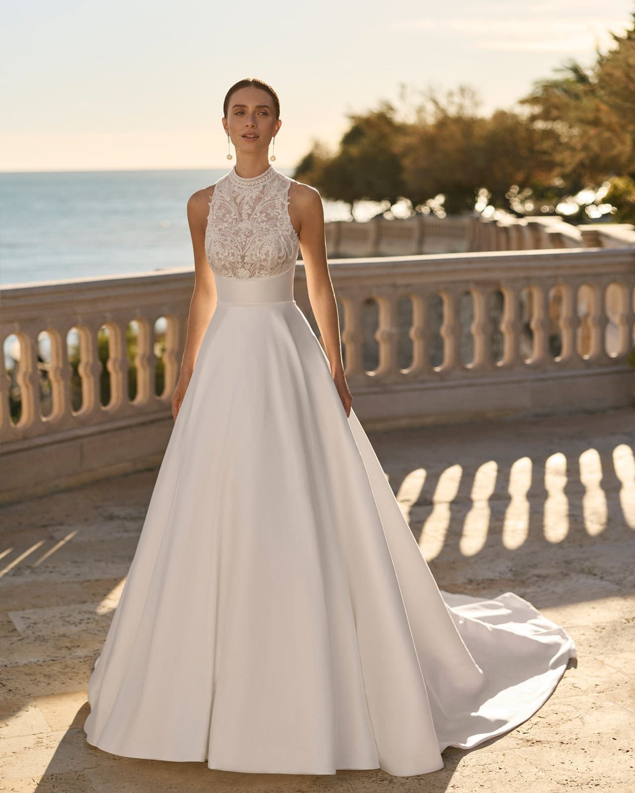 Mujer con un vestido de gala de encaje blanco de pie en una terraza junto al mar al atardecer.