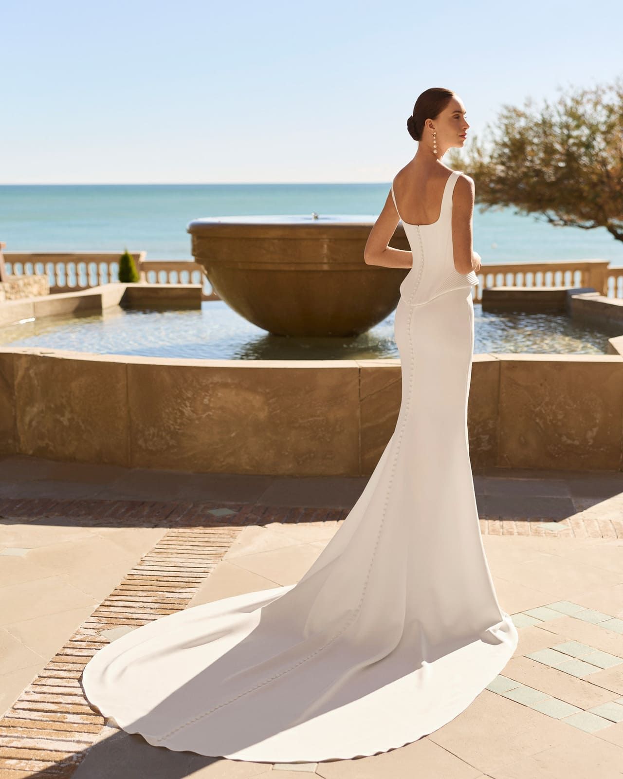 Mujer con un vestido blanco de pie junto a una fuente junto al mar, con una larga cola que se arrastra por el patio.
