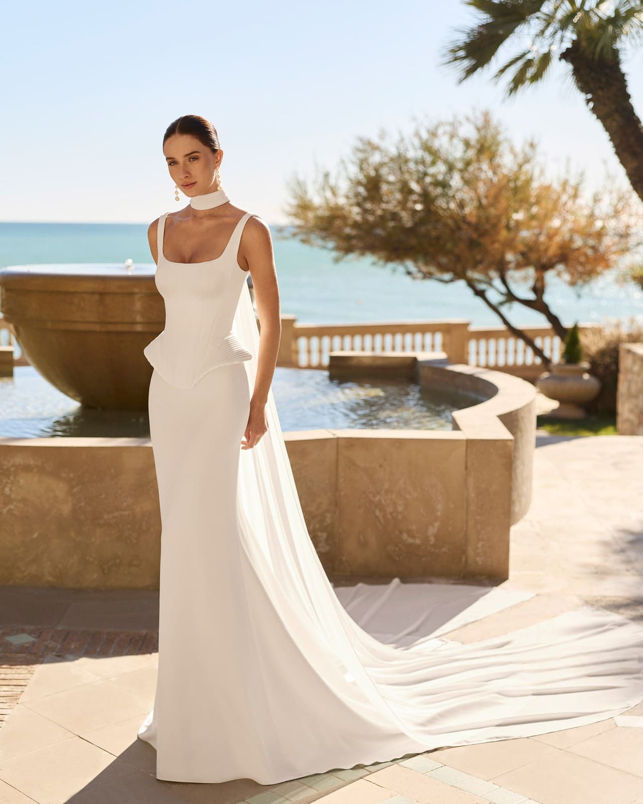 Mujer con vestido blanco de pie al aire libre junto a una piscina con vista al mar y palmeras.