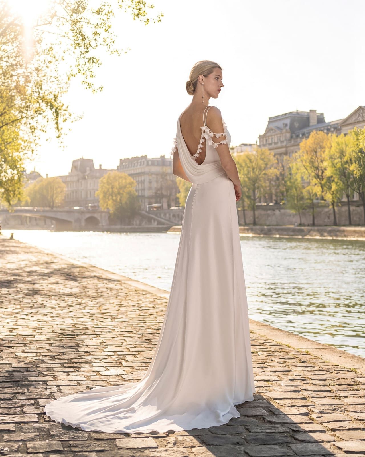 Mujer con un vaporoso vestido blanco de pie junto a un río en un sendero empedrado iluminado por el sol.