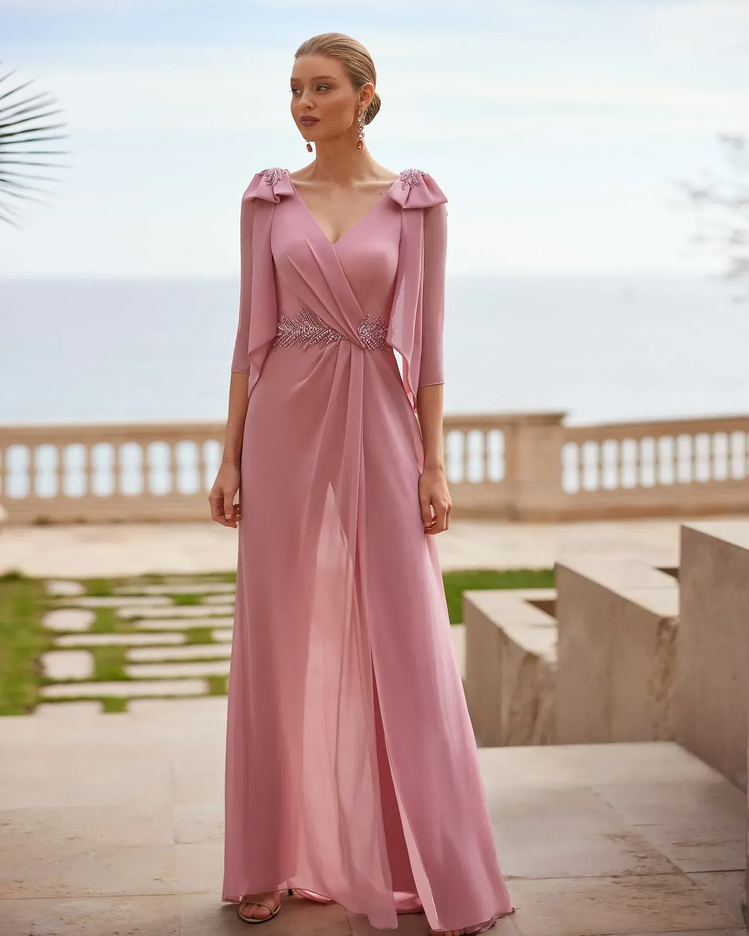Una mujer con un vestido largo de color rosa está parada en las escaleras frente al océano.