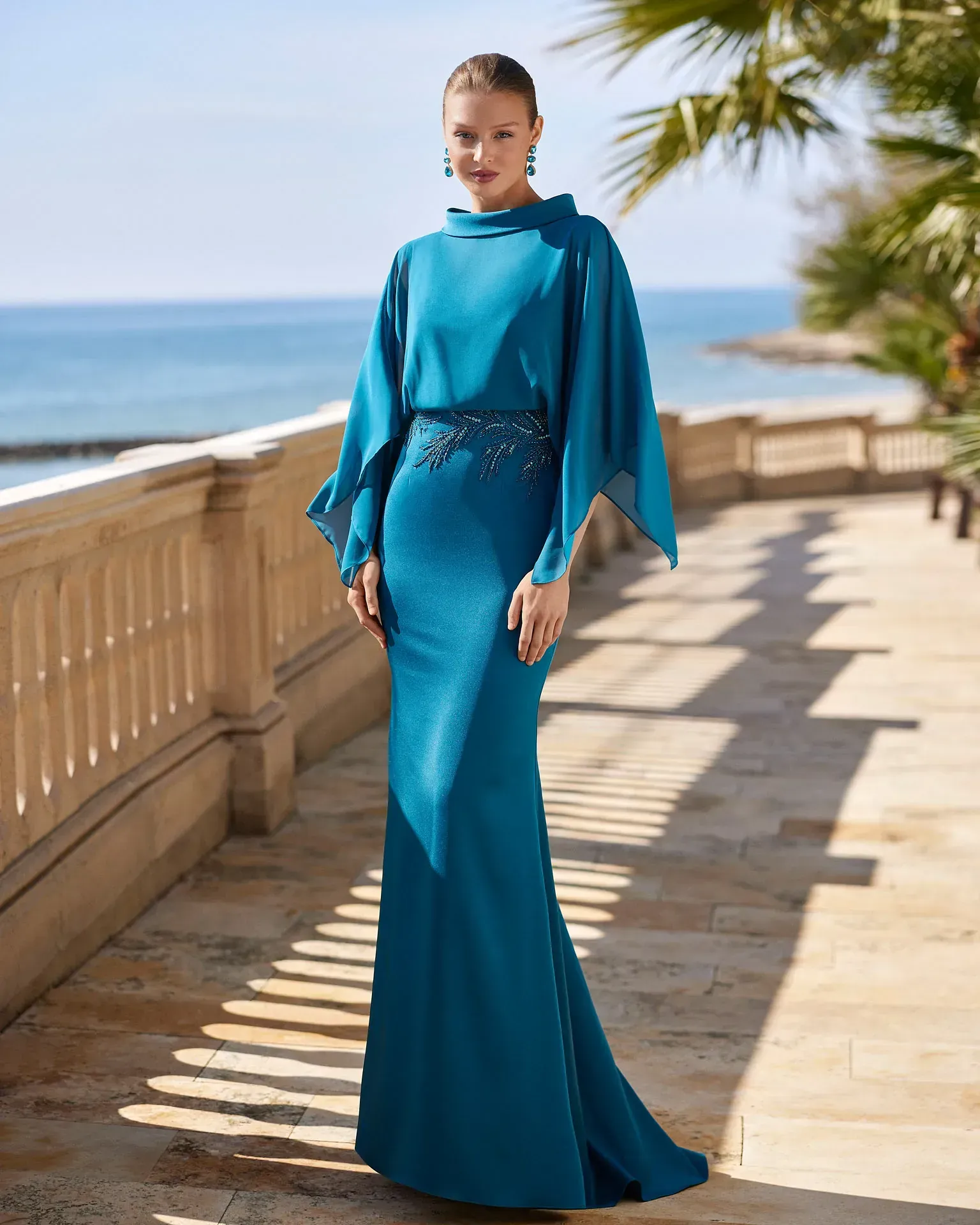 Una mujer con un vestido largo azul está parada en un balcón con vista al océano.