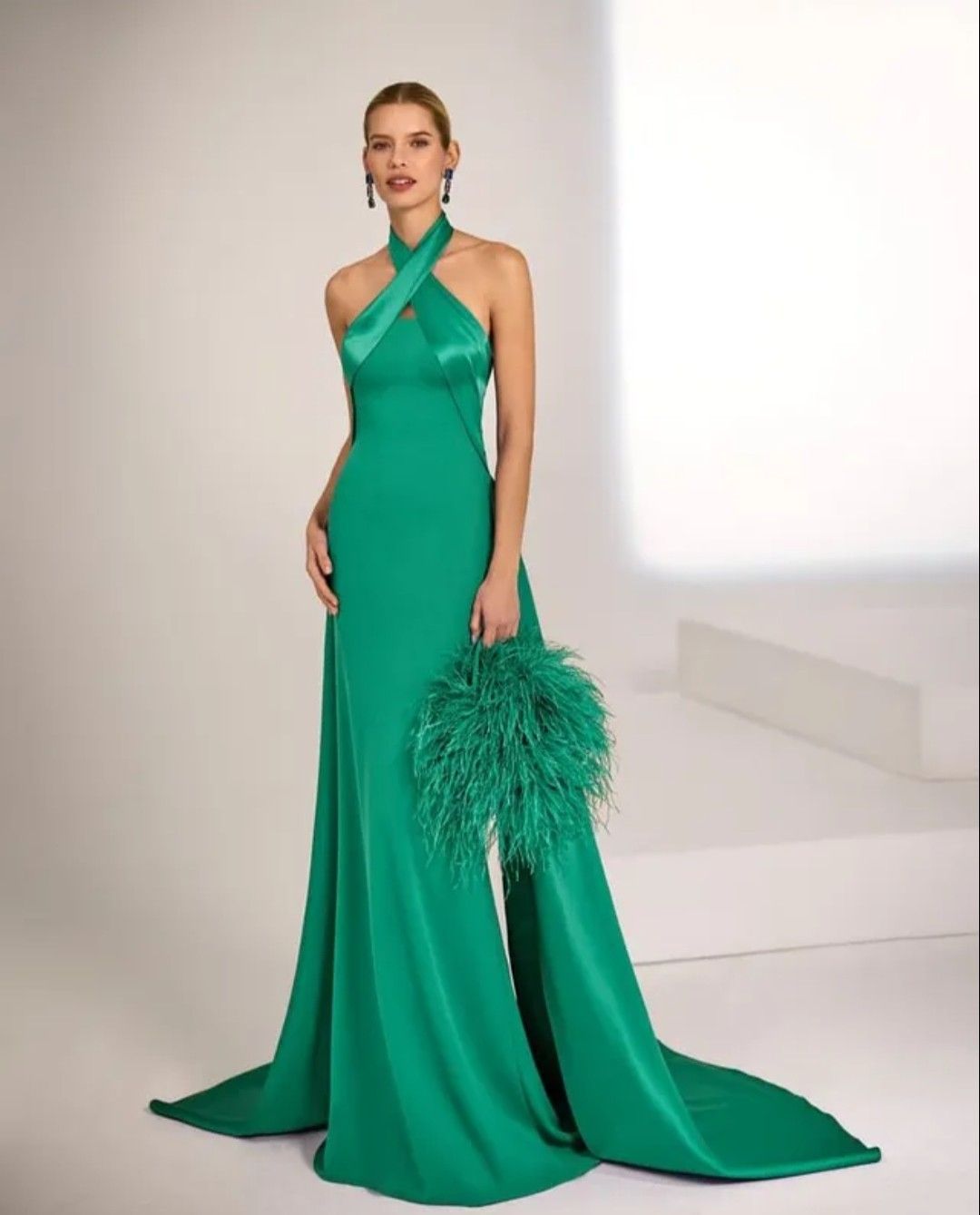 Mujer con vestido verde sosteniendo un bolso de mano con plumas en una pasarela blanca.