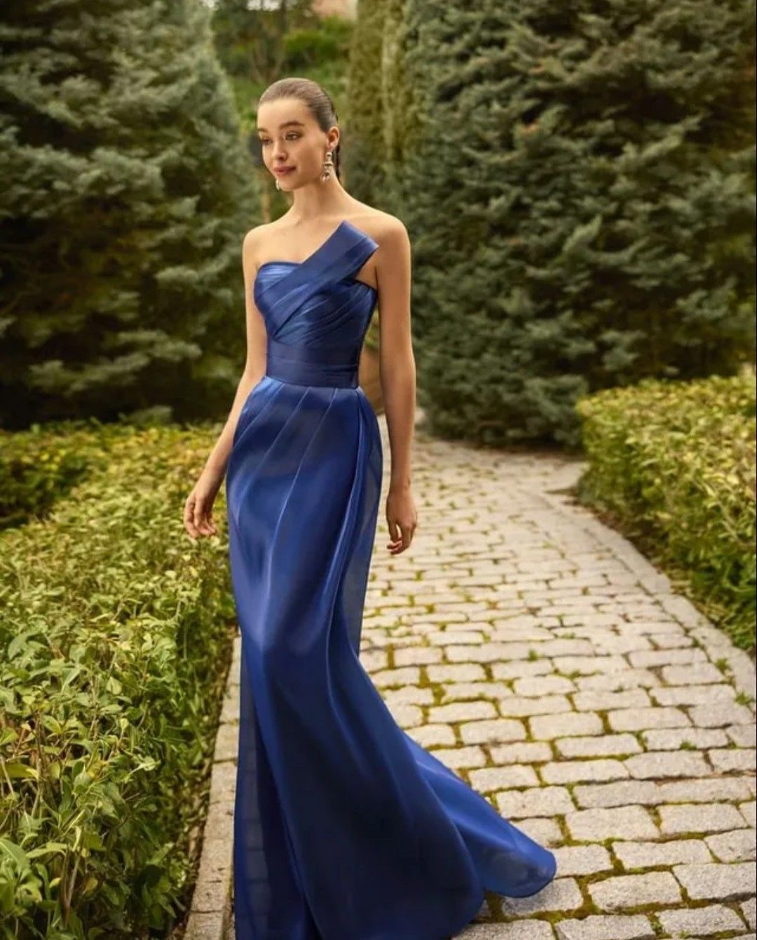 Mujer con un vaporoso vestido azul real de pie sobre un sendero empedrado en un jardín.