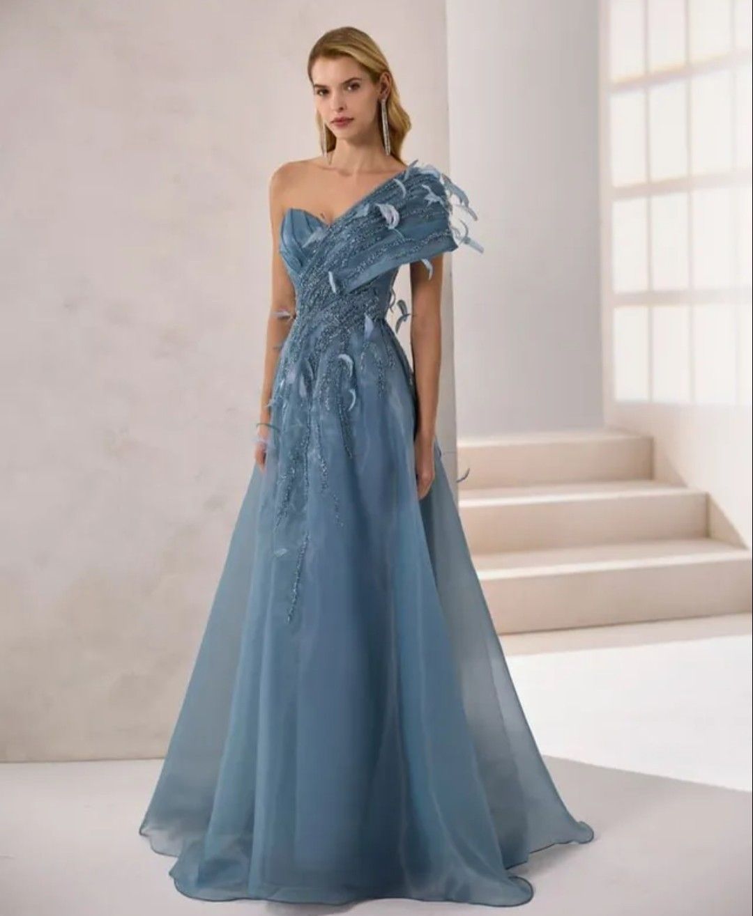 Vestido de noche azul de un solo hombro con apliques florales, lucido por una modelo en una escalera.