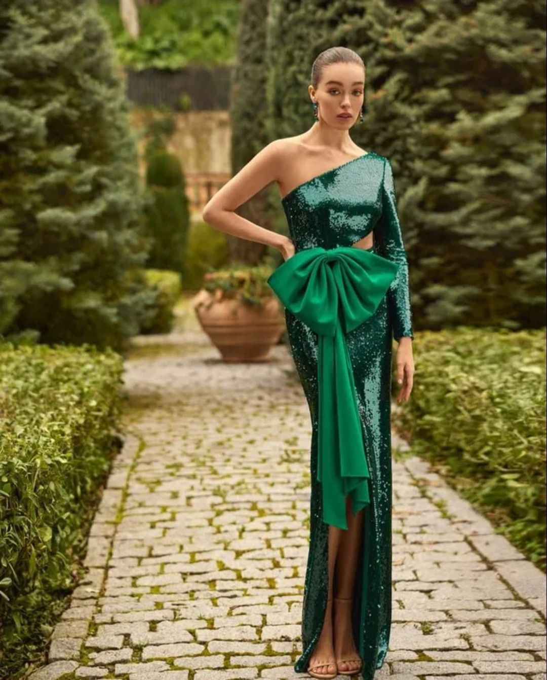 Mujer con un vestido de satén verde posando en un sendero de jardín entre setos y macetas.
