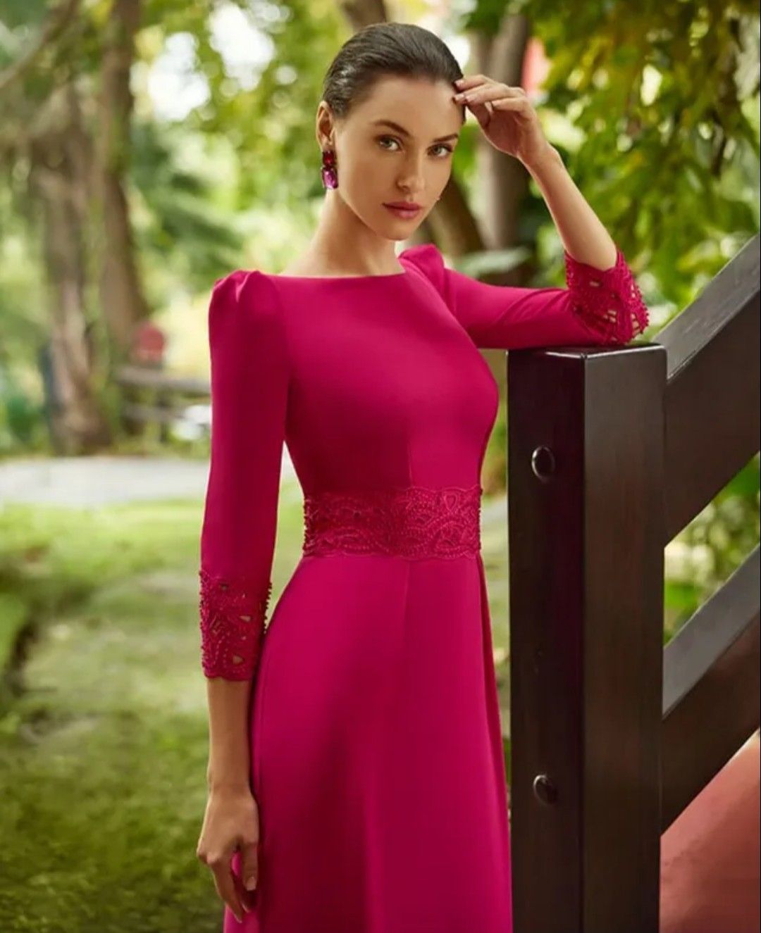 Mujer con un vestido rosa brillante de pie junto a una barandilla de madera al aire libre, con la mano levantada hacia su cabello.