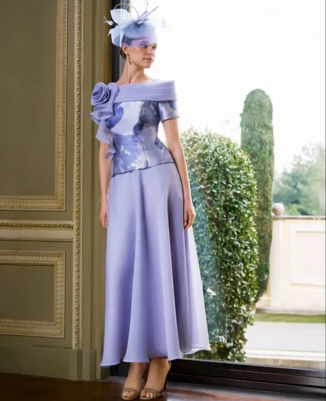 Vestido lavanda con los hombros descubiertos y sombrero a juego en una modelo de pie junto a una puerta con vista al jardín.