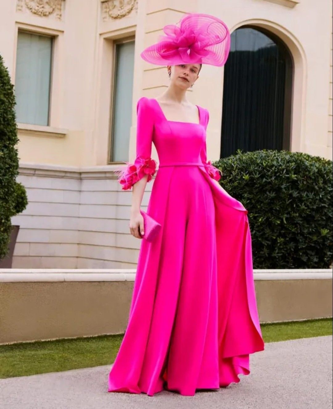 Mujer con un vestido rosa brillante y sombrero a juego de pie al aire libre junto a un edificio.