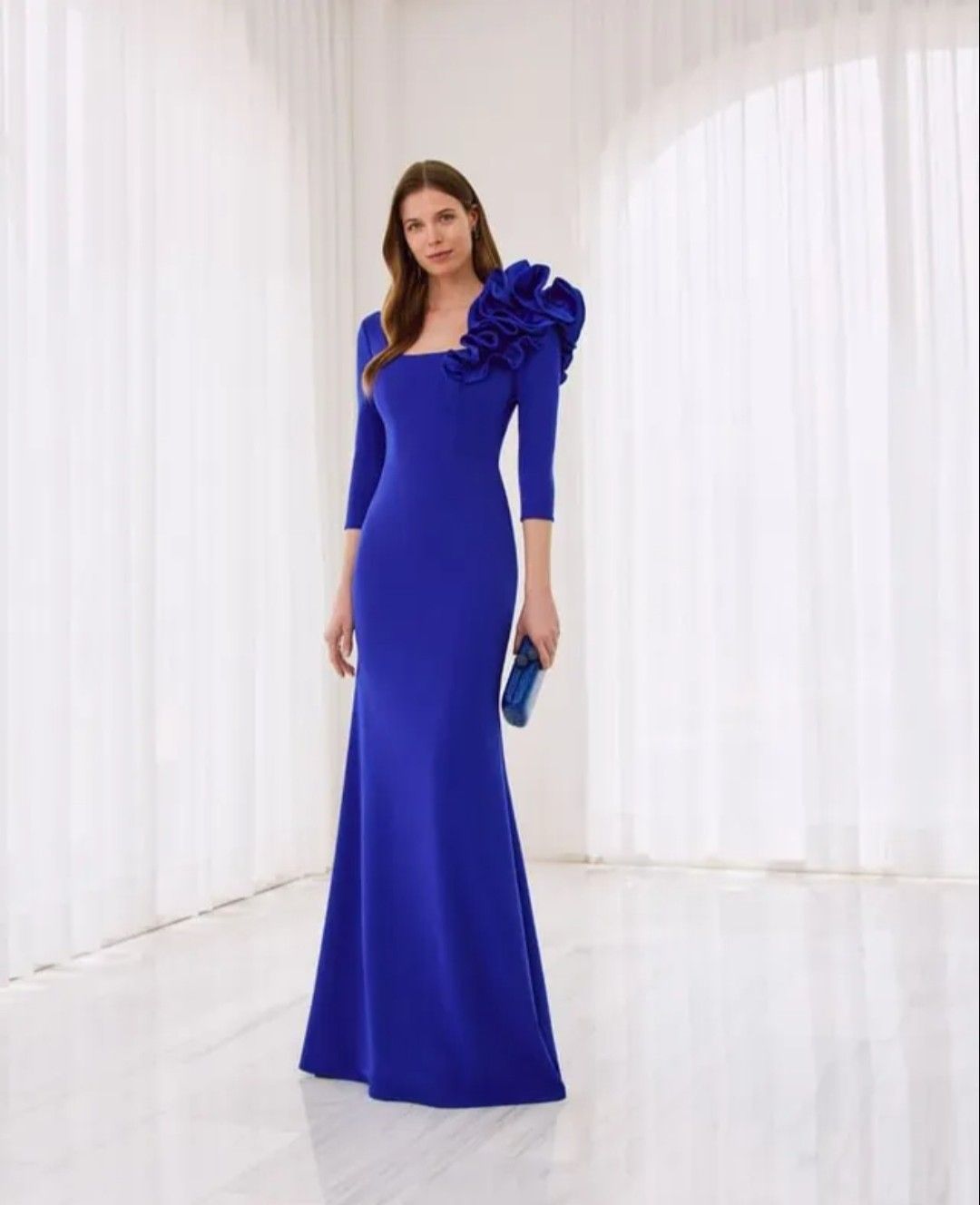 Mujer con un vestido largo azul marino con detalles florales en los hombros, de pie en una habitación blanca luminosa.