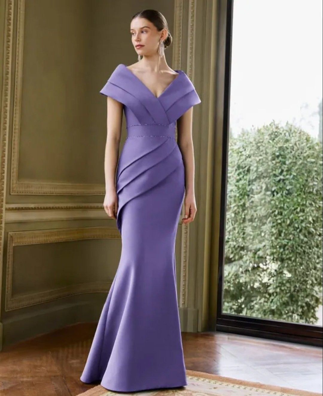 Mujer con un vestido de noche lila con los hombros descubiertos de pie junto a una gran ventana en una sala formal.
