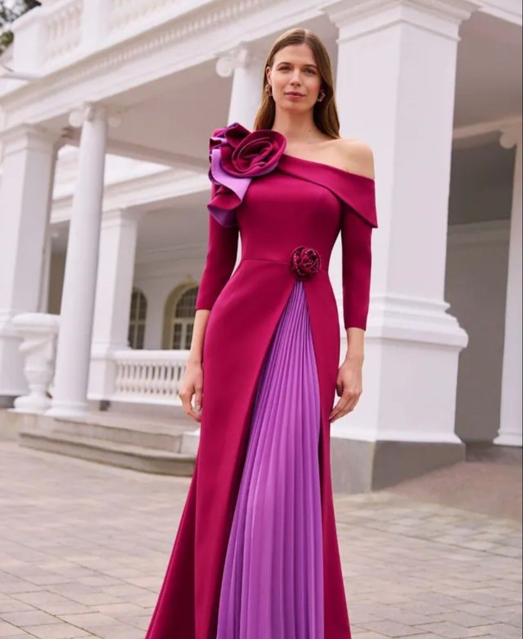 Mujer con un vestido magenta y morado de pie en un patio con columnas blancas.