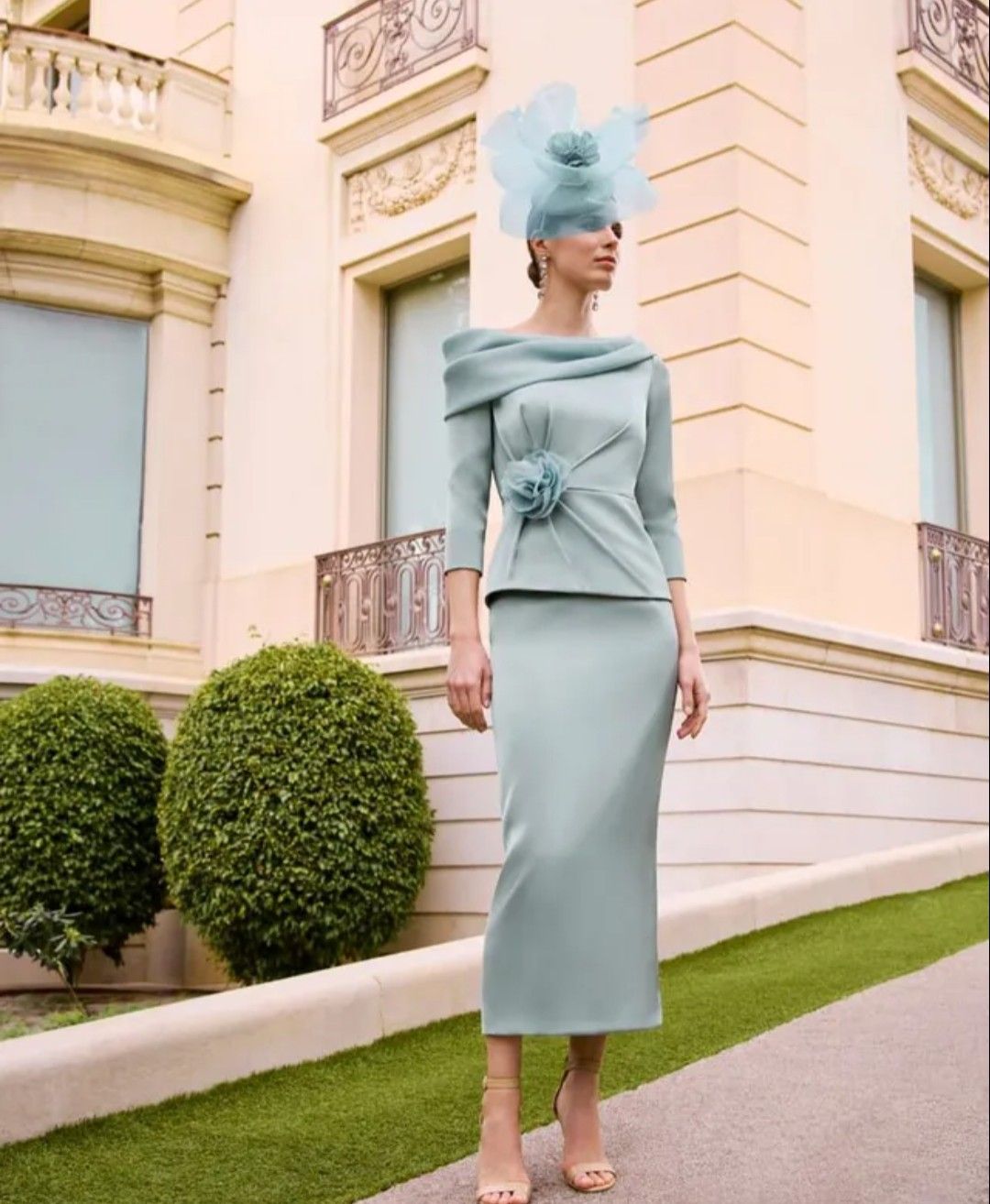 Mujer con vestido y sombrero azul claro posando frente a un edificio de tonos pastel y arbustos bien cuidados.