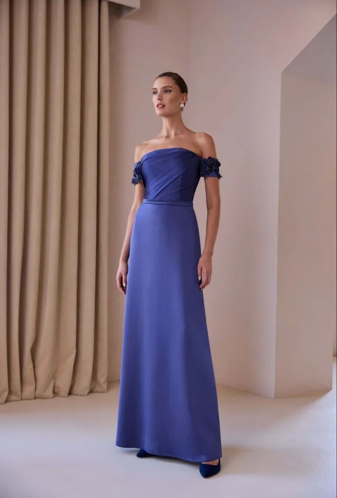 Mujer con un vestido azul sin tirantes de pie en una habitación minimalista con cortinas beige.