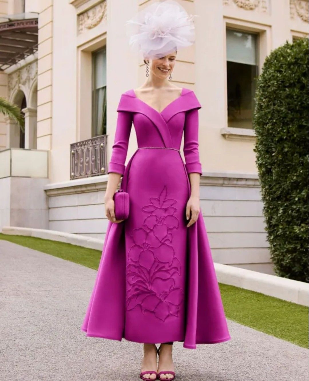 Mujer con vestido y sombrero de color magenta brillante posando al aire libre junto a un edificio y setos podados.