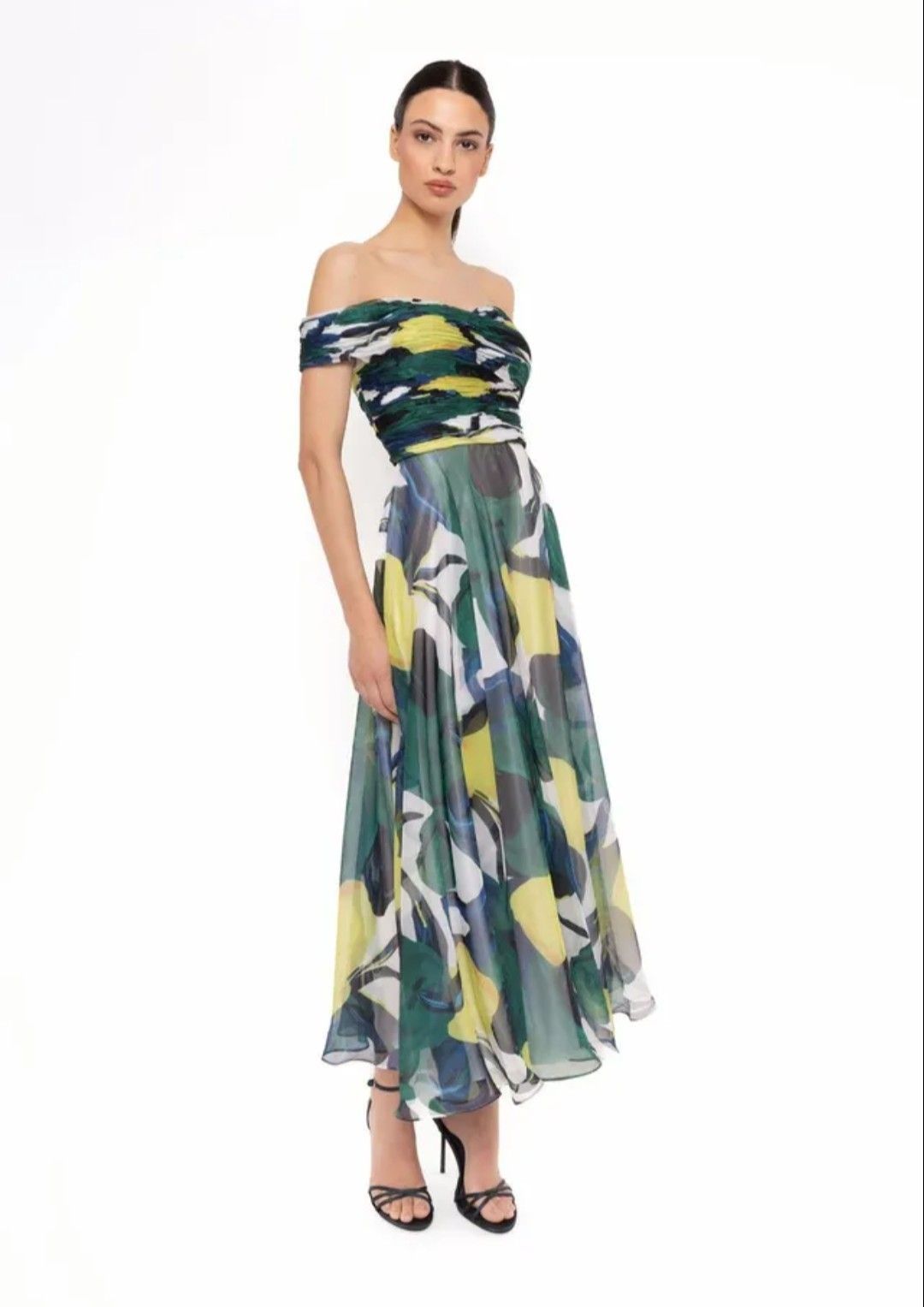 Mujer con vestido largo floral de hombros descubiertos, falda transparente y tacones negros sobre fondo blanco.