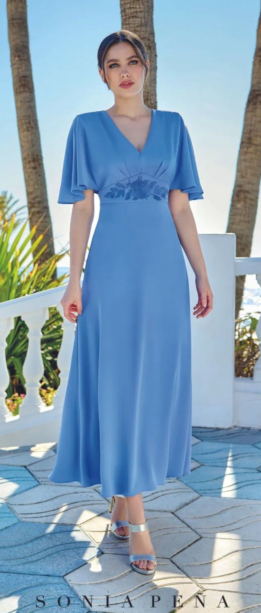 Mujer con un vestido largo azul claro de pie en una terraza soleada junto al mar.