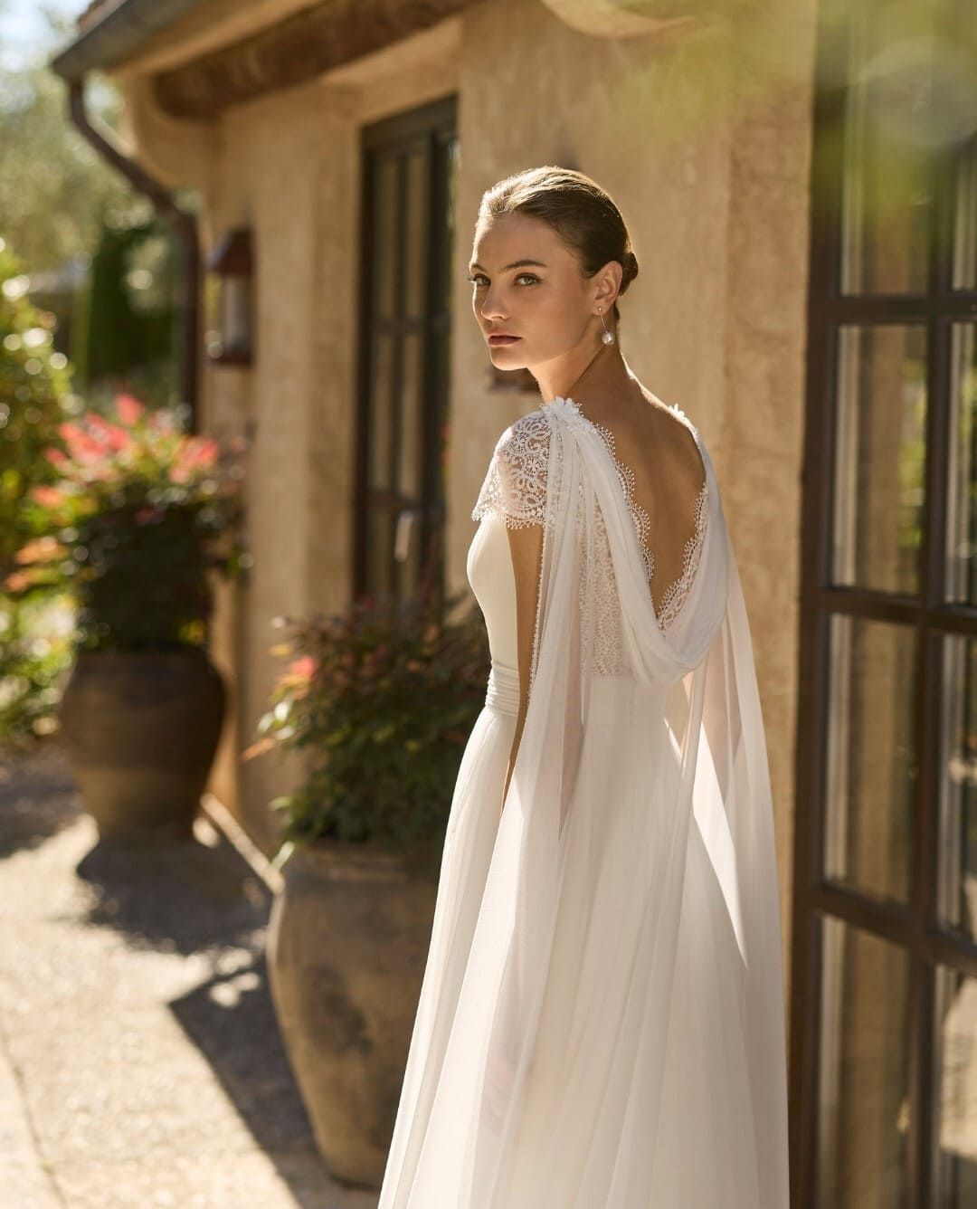 Novia con un vestido de encaje blanco de pie al aire libre junto a un muro de piedra iluminado por el sol y macetas con plantas.