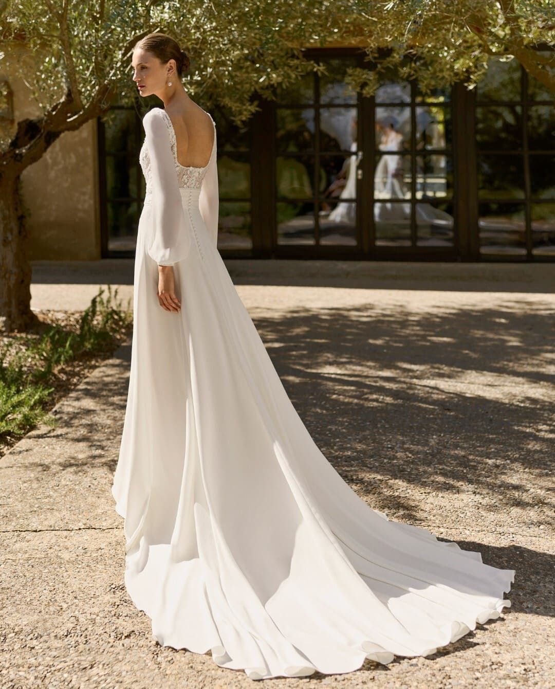 Mujer con un vestido de novia blanco con una larga cola de pie al aire libre junto a un edificio y árboles.