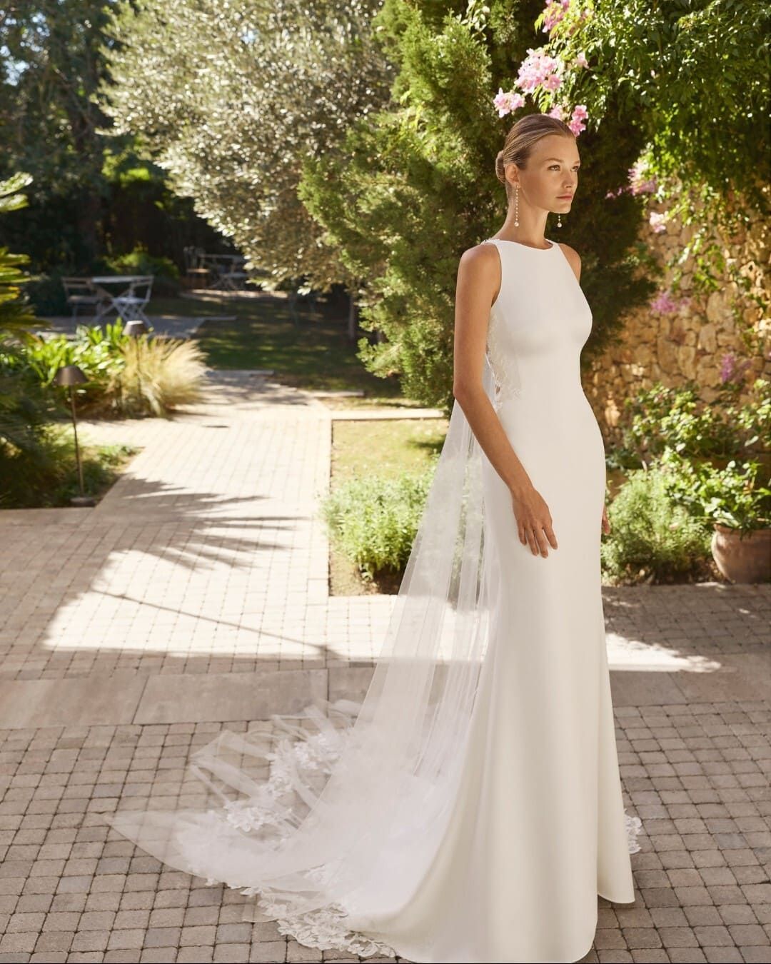 Novia con vestido blanco y velo largo, de pie en un sendero soleado de un jardín.