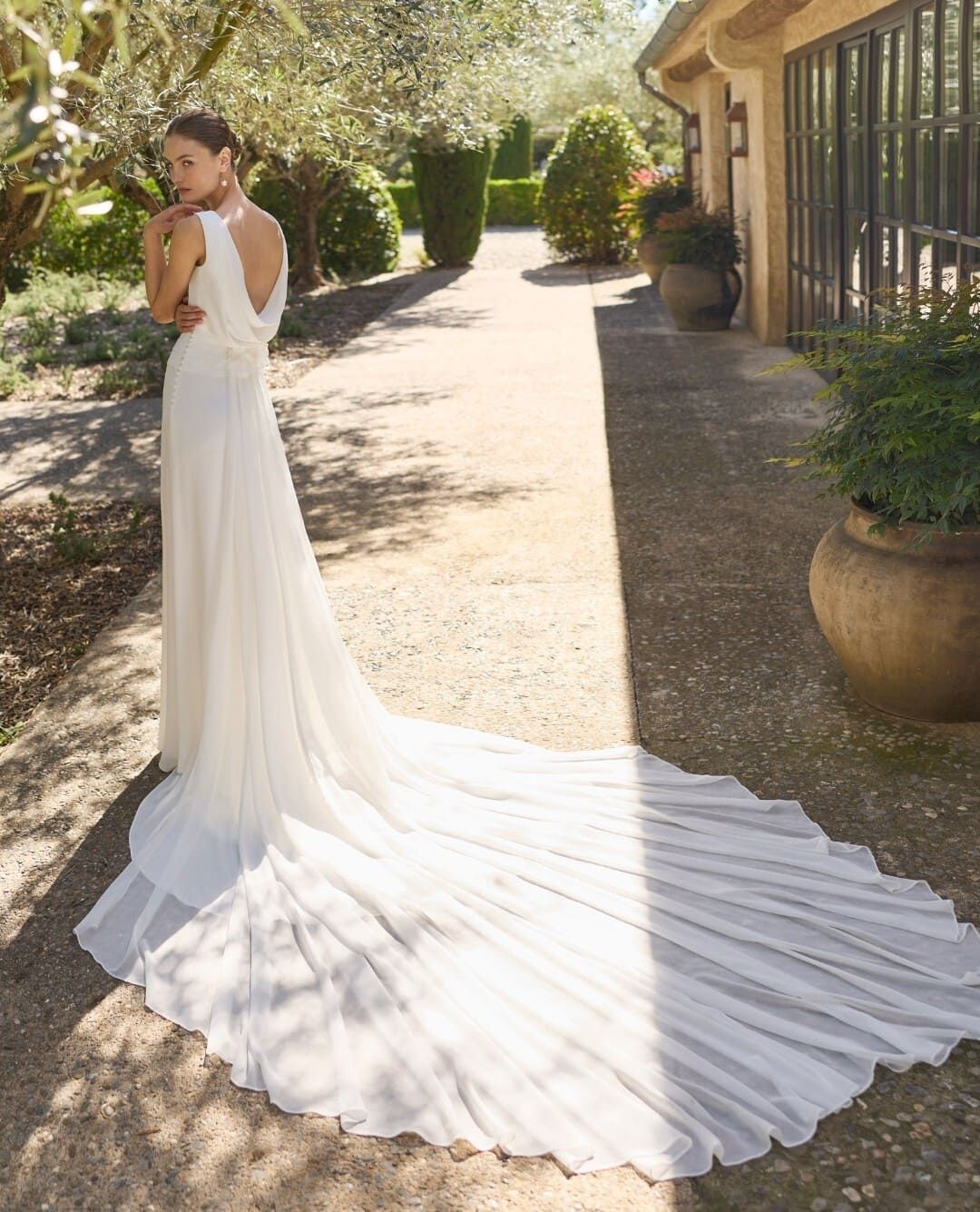 Novia con un vestido blanco de larga cola de pie en un sendero de jardín iluminado por el sol.