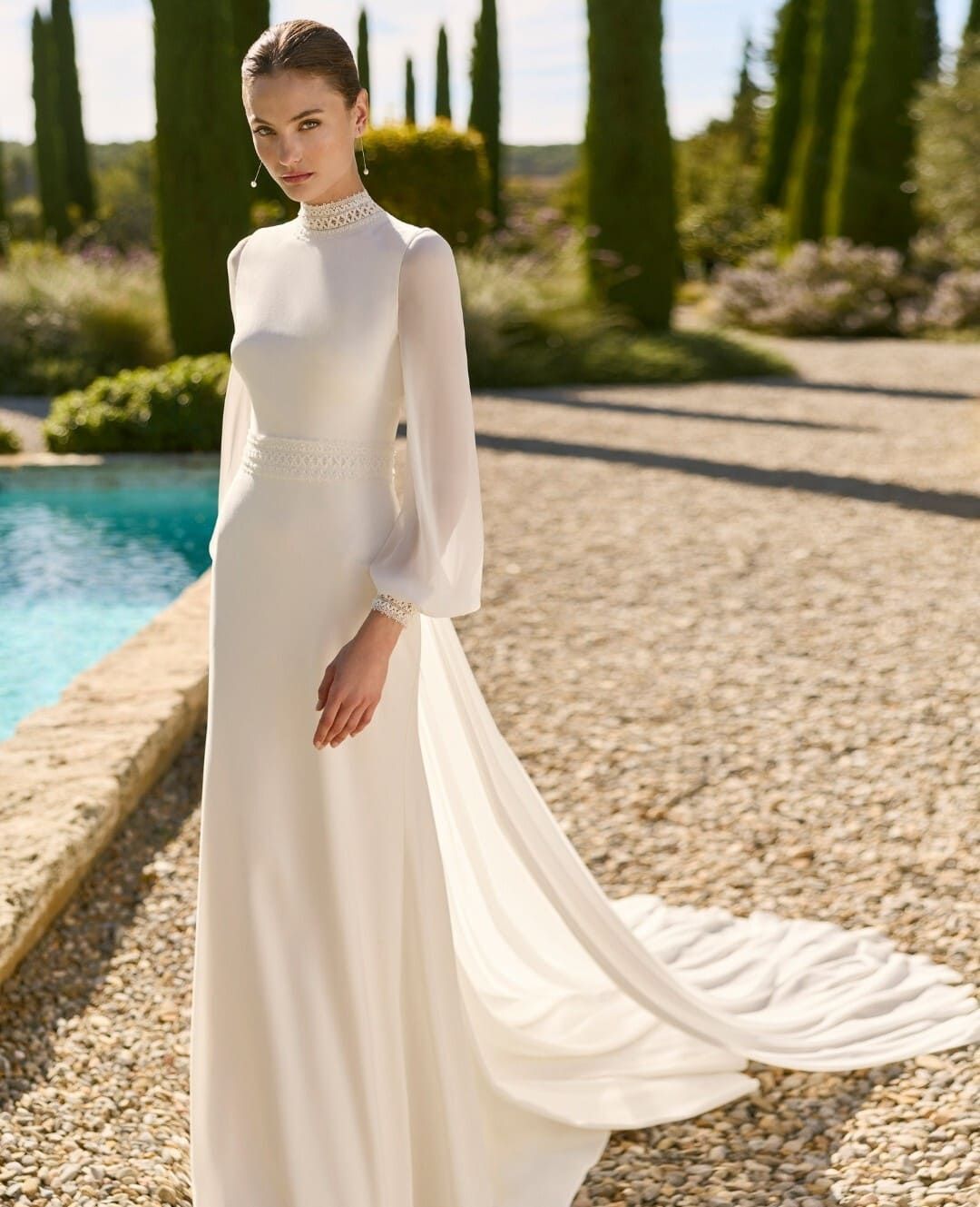 Mujer con un largo vestido blanco de pie junto a una piscina turquesa en un sendero de jardín iluminado por el sol.