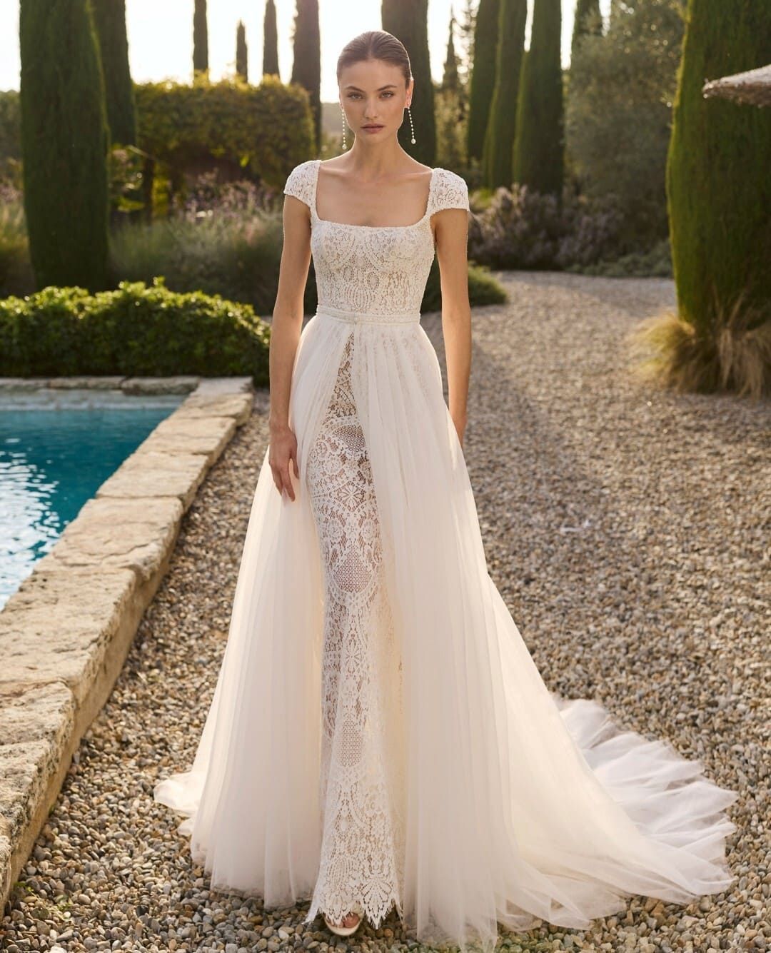 Modelo con un vestido de novia de encaje blanco de pie junto a una piscina en un entorno ajardinado.