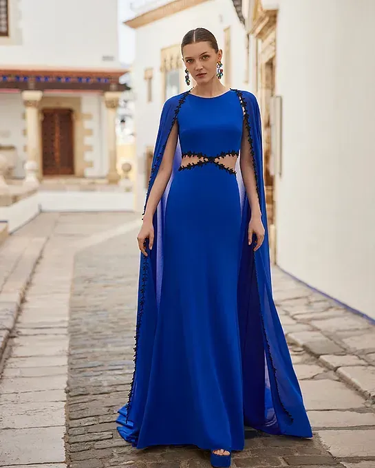 Una mujer con un vestido largo azul y una capa está parada en una acera.