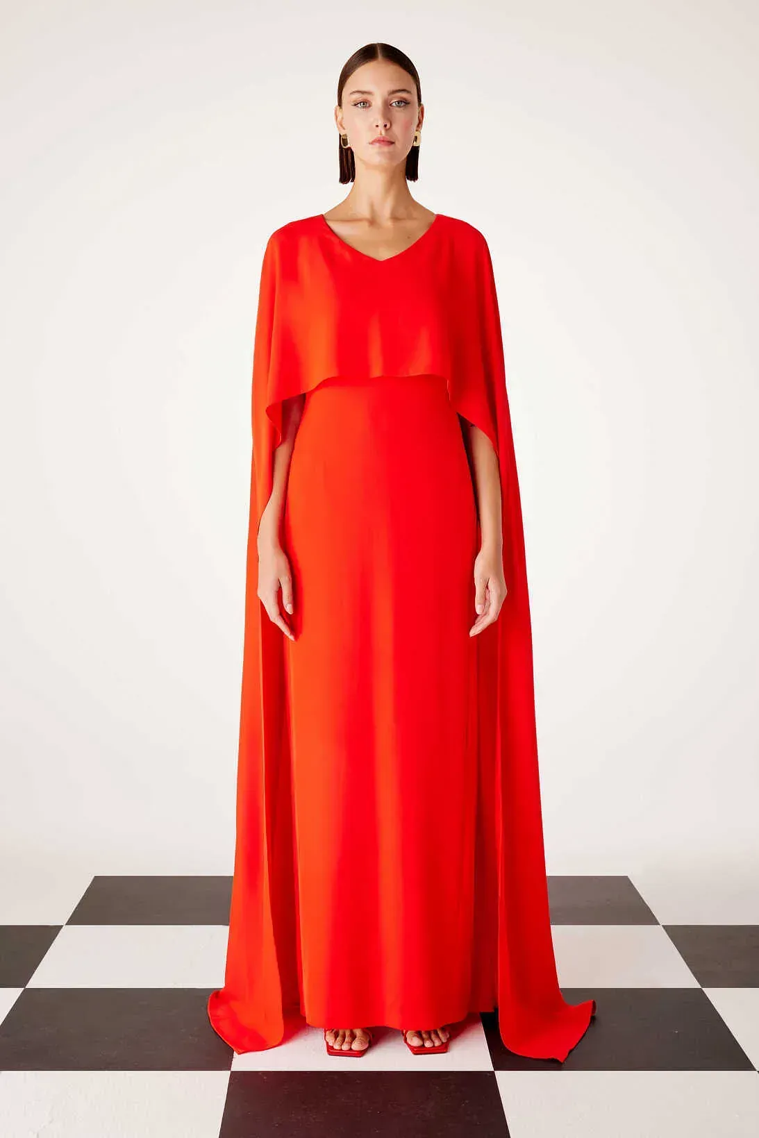 Una mujer con un vestido rojo y una capa está parada sobre un piso a cuadros.