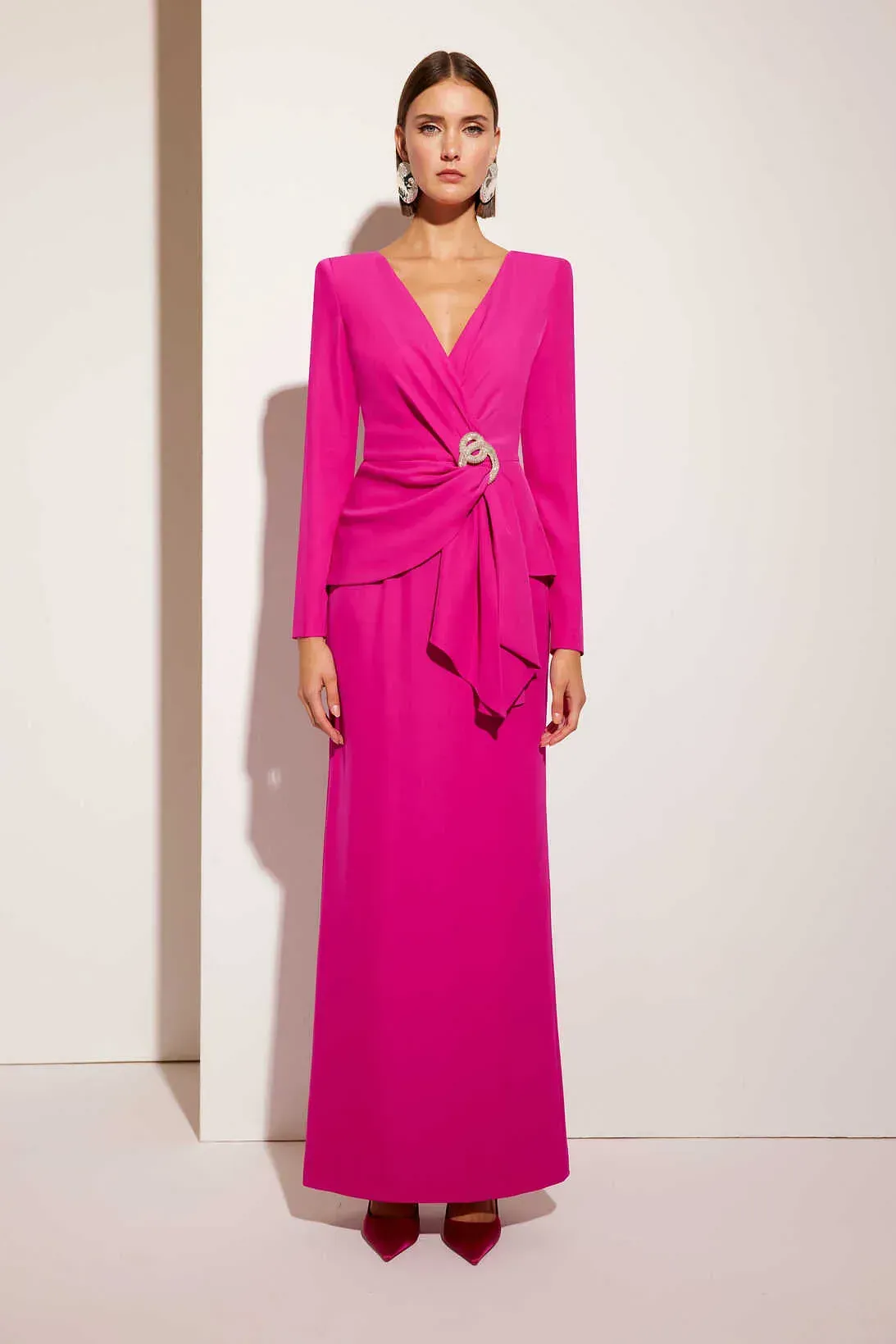 Una mujer con un vestido largo de color rosa está parada frente a una pared blanca.