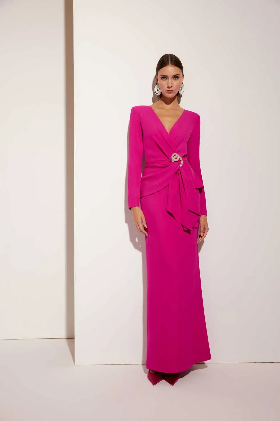 Una mujer con un vestido largo de color rosa está parada frente a una pared blanca.
