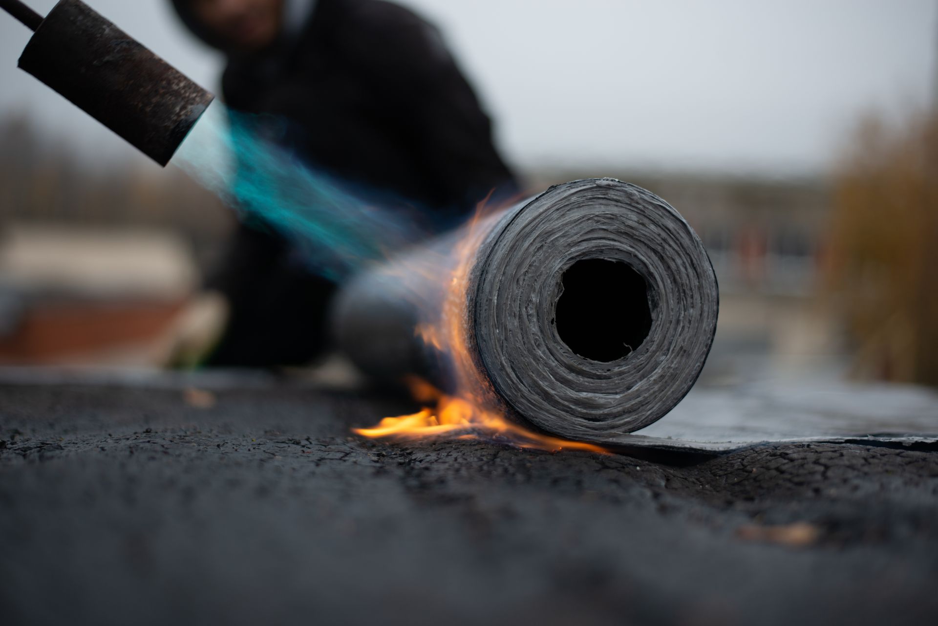 Un ouvrier utilise un chalumeau à propane pour faire fondre un rouleau de bitume noir sur une surface plane.