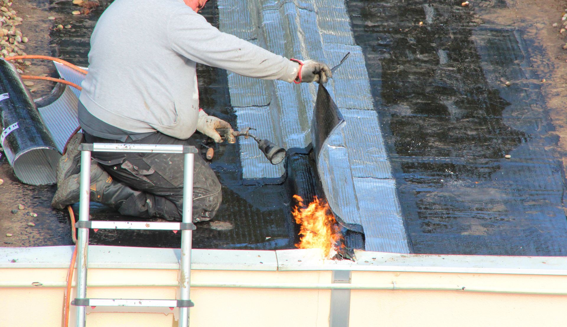 Un ouvrier utilise un chalumeau à propane pour installer une membrane d'étanchéité, la faisant fondre sur la surface d'un toit plat.