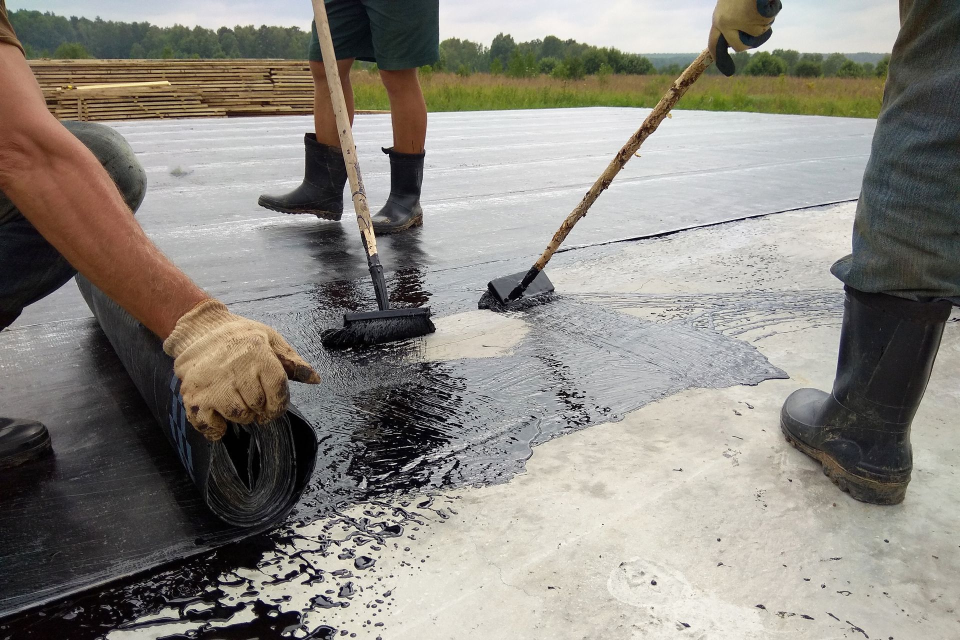 Des ouvriers appliquent un adhésif bitumineux noir sur une surface en béton à l'aide de pinceaux à long manche tout en déroulant le matériau de toiture.