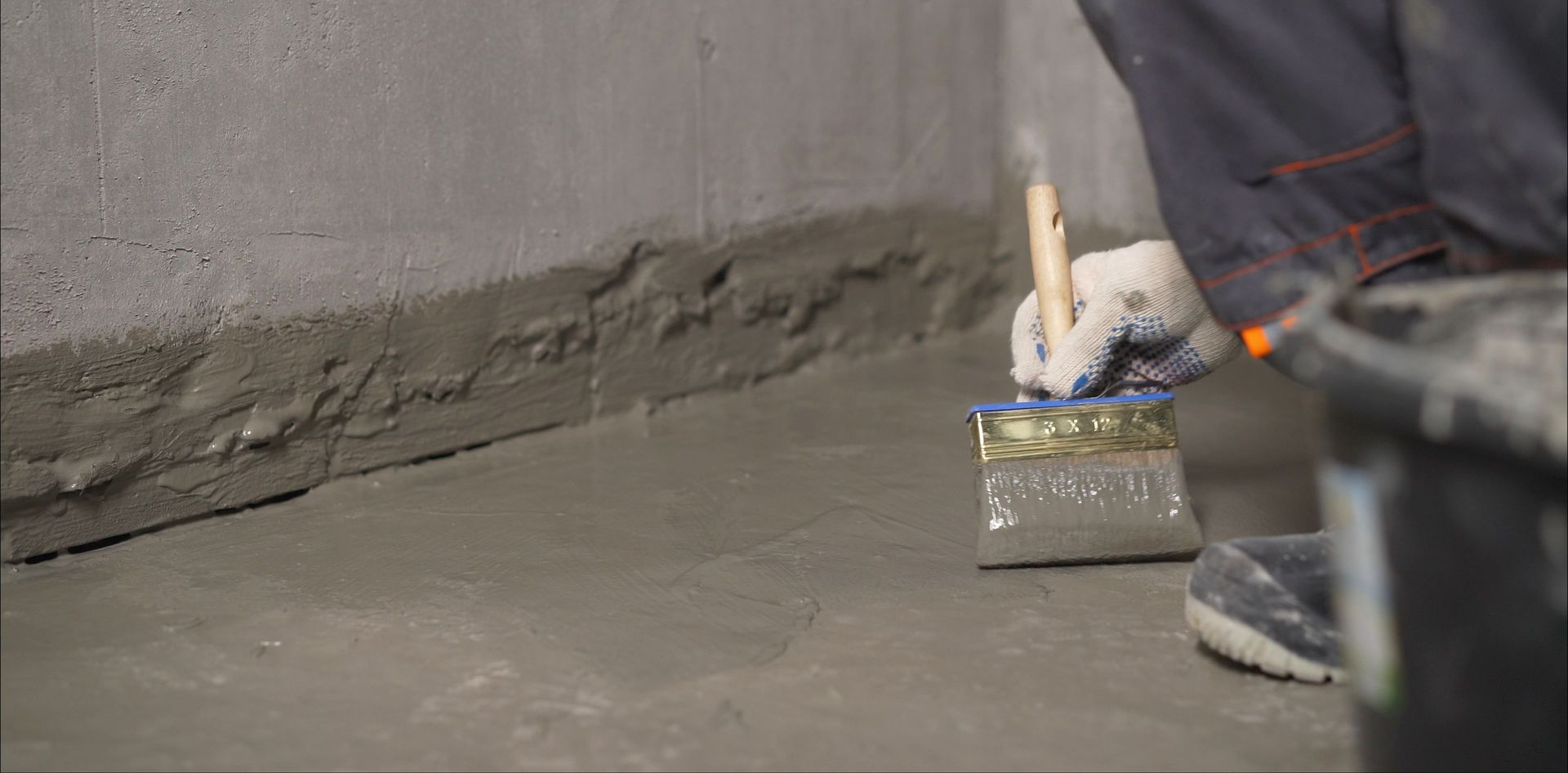 Une personne utilise un large pinceau pour appliquer un mastic d'étanchéité gris imperméable sur le joint entre un mur et un sol.