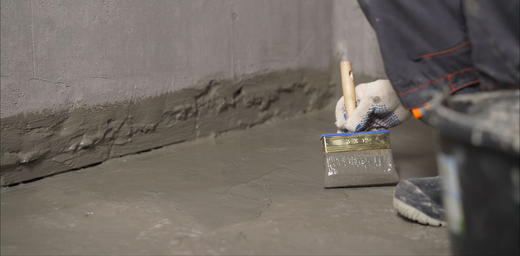 Une personne utilise un large pinceau pour appliquer un mastic d'étanchéité gris imperméable sur le joint entre un mur et un sol.