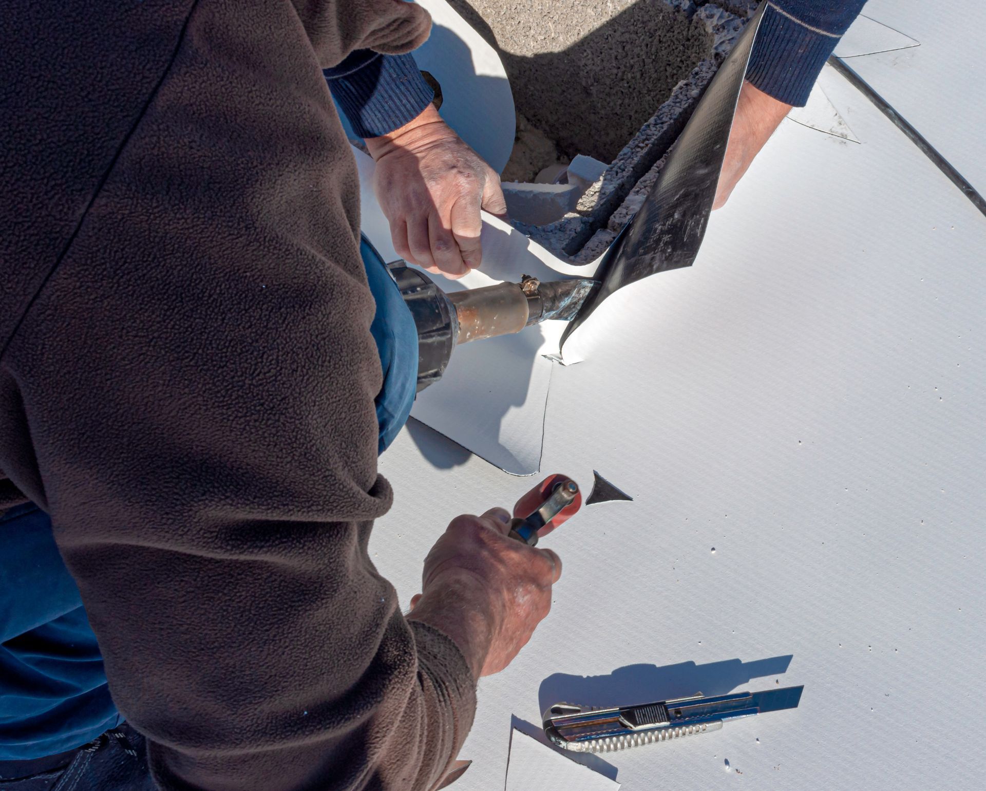 Deux ouvriers utilisent un pistolet thermique et un outil manuel pour souder les joints d'une membrane de toiture blanche sur un toit plat.