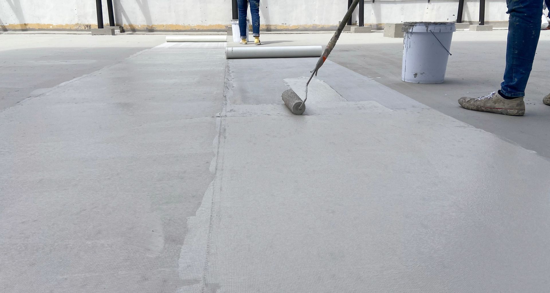 Un ouvrier utilise un rouleau à peinture pour appliquer un mastic d'étanchéité liquide gris sur la surface plane d'un toit en béton.