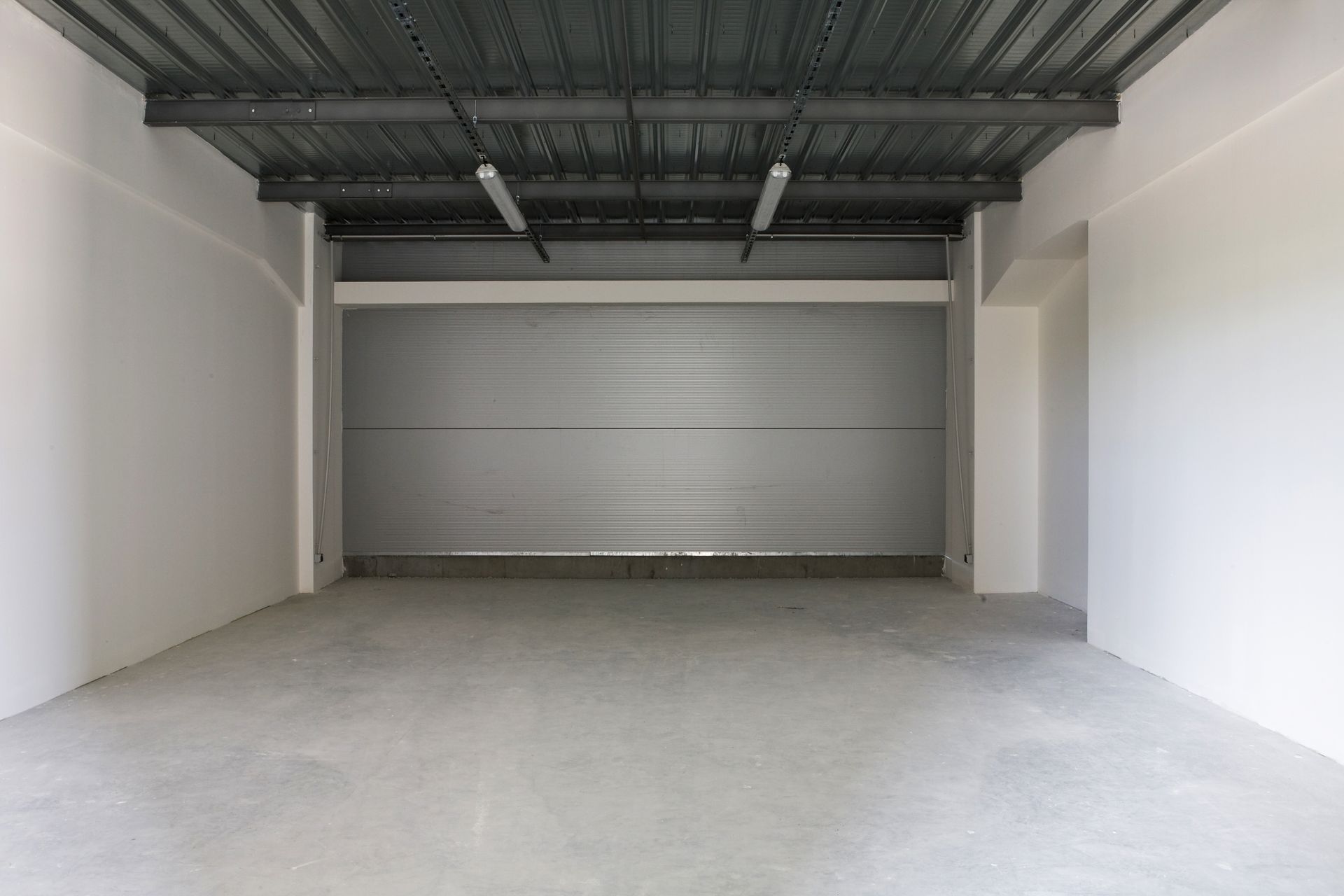 Une pièce vide, de style industriel, avec des murs blancs, un sol en béton gris et un plafond en tôle ondulée sombre.