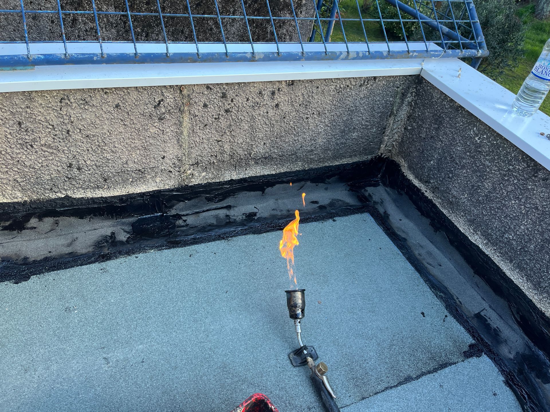 Un ouvrier portant un casque de chantier et un pantalon bleu utilise un chalumeau à propane pour faire fondre un matériau de toiture bitumineux sur une surface.