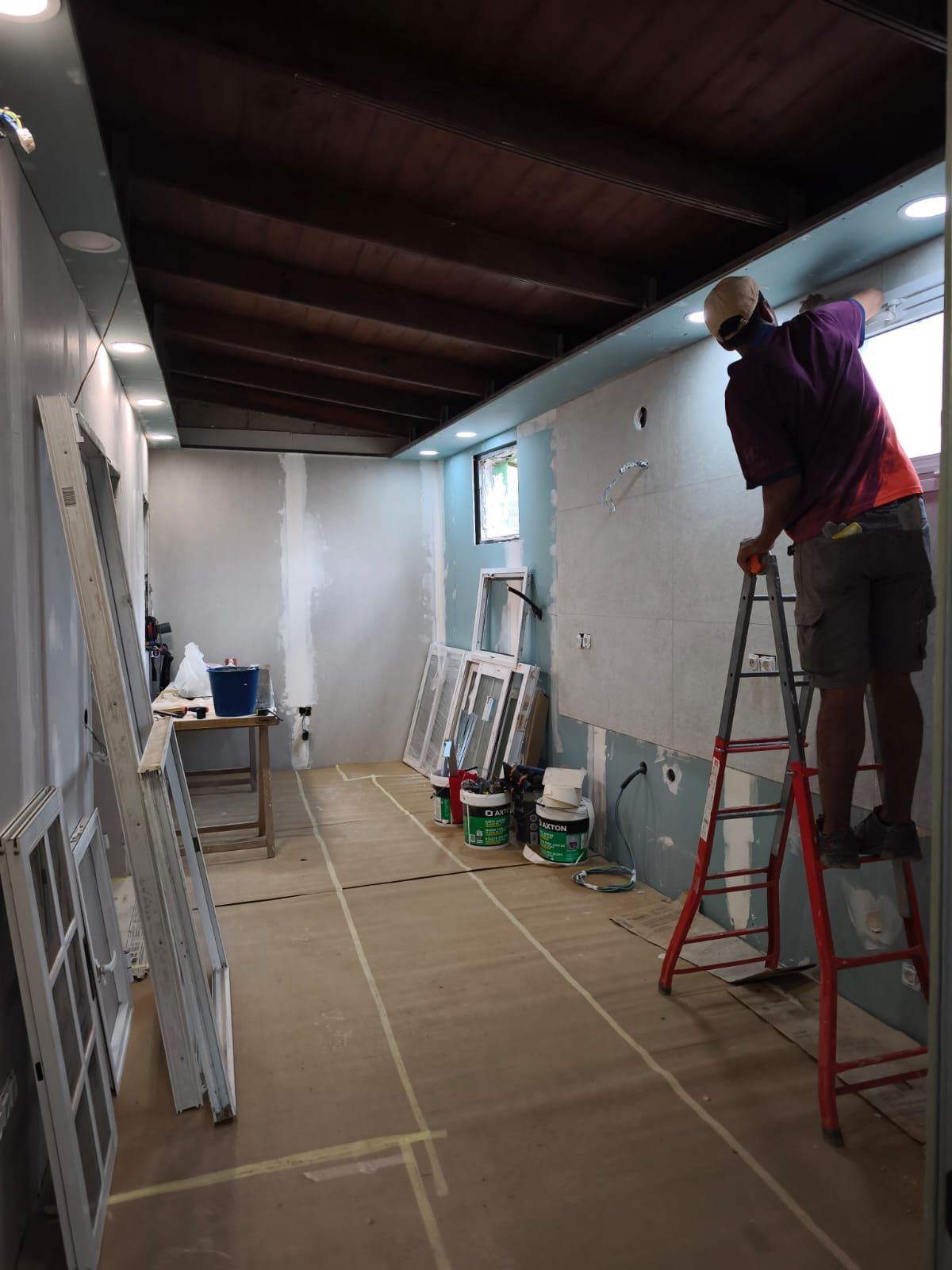 Un trabajador subido a una escalera pinta una habitación estrecha con paredes blancas y un techo de madera oscura.