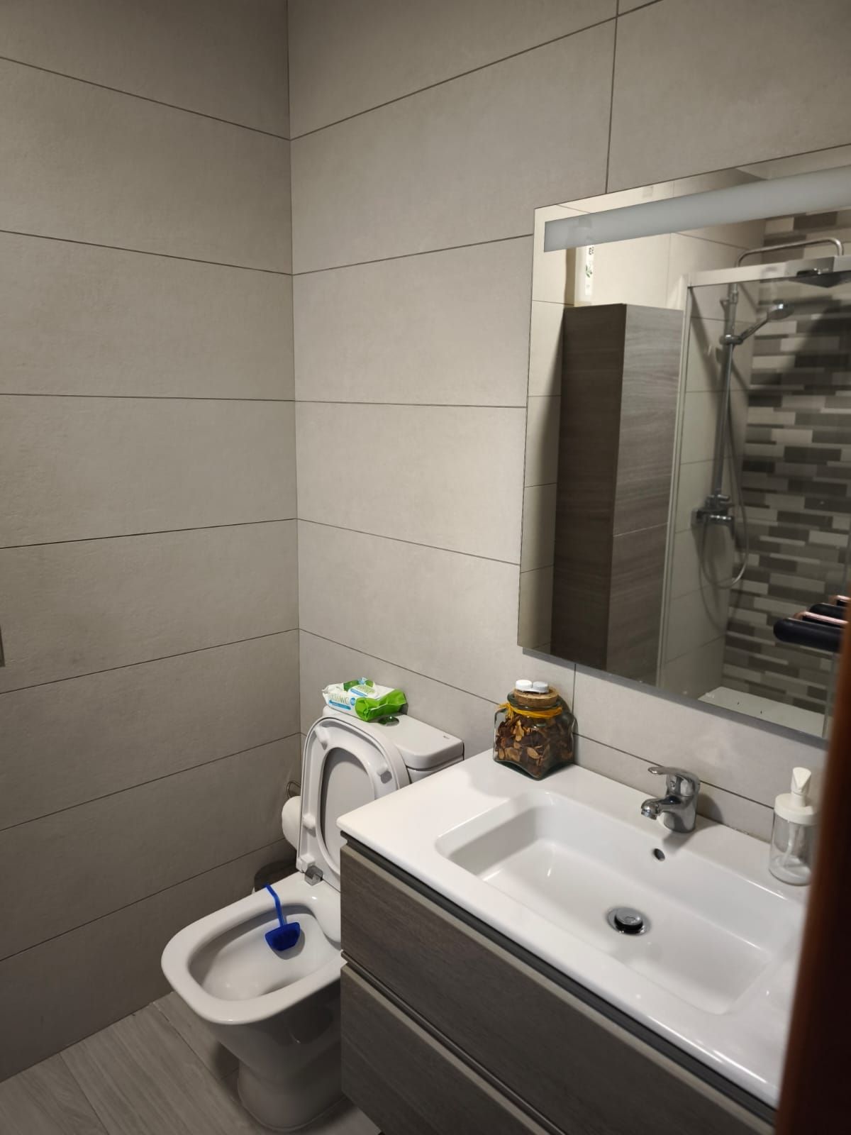 Baño pequeño con inodoro, lavabo, espejo y paredes revestidas de azulejos grises.