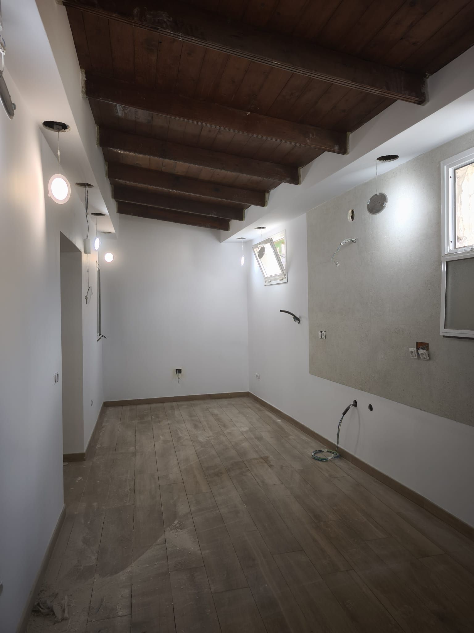 Habitación vacía y sin amueblar, con paredes blancas, vigas de madera en el techo y suelo gris, en obras.
